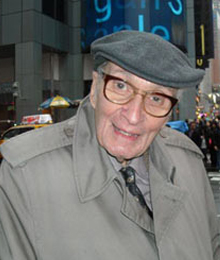 John Balamos smiling on a New York City street, wearing a cap and overcoat